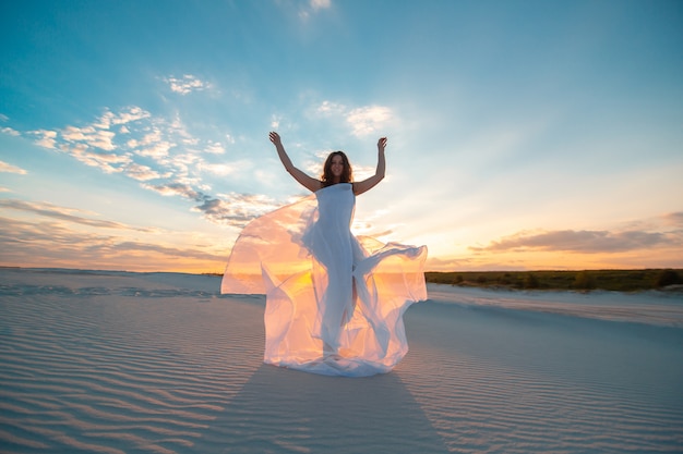 A girl in a fly white dress dances and poses in the sand desert at sunset