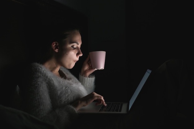 girl freelancer sitting in bed late at night, working on laptop and drinking coffee