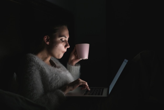 Girl freelancer sitting in bed late at night, working on laptop and drinking coffee