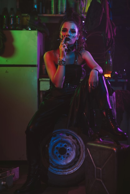 Girl in a futuristic cyberpunk costume smokes a cigarette with a neon light in the garage. Steampunk style. Futuristic post-apocalyptic style