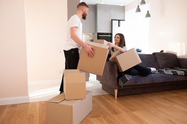 Girl giving cardboard box with things to her boyfriend at home. Joyful european couple of woman and man. Concept of moving in new flat. Idea of young family. Interior of studio apartment. Sunny day
