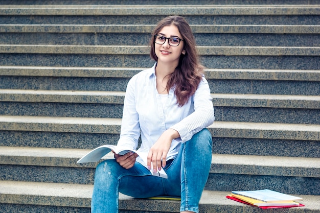 Una ragazza con gli occhiali con un libro si siede sui gradini