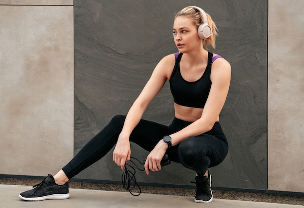 Girl goes in for sports on the street, Black clothing for sports, Woman in sports tops, Girl in headphones, Girl sitting near the wall