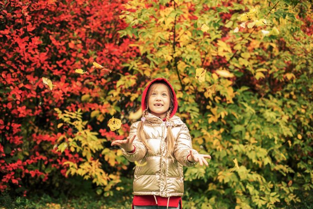 Foto una ragazza con una giacca color oro sorride lanciando foglie rosa e gialle in una giornata autunnale