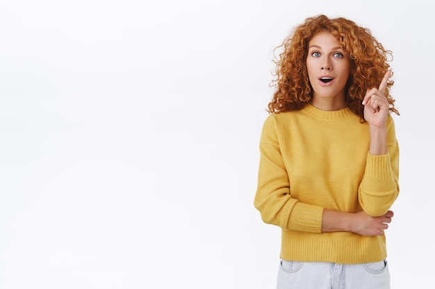 Foto la ragazza ha avuto un'idea eccellente, aggiungendo un suggerimento. donna rossa con i capelli ricci in maglione giallo, alza un dito nel gesto di eureka