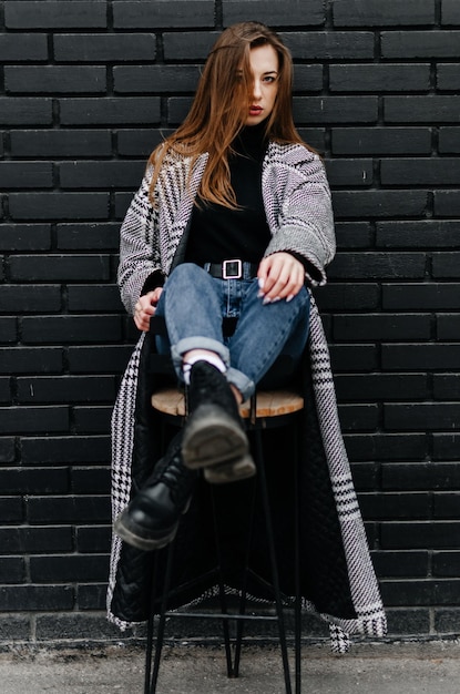 a girl in a gray coat sits on a chair near a black wall