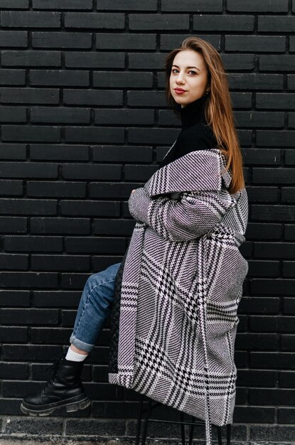 a girl in a gray coat sits on a chair near a black wall