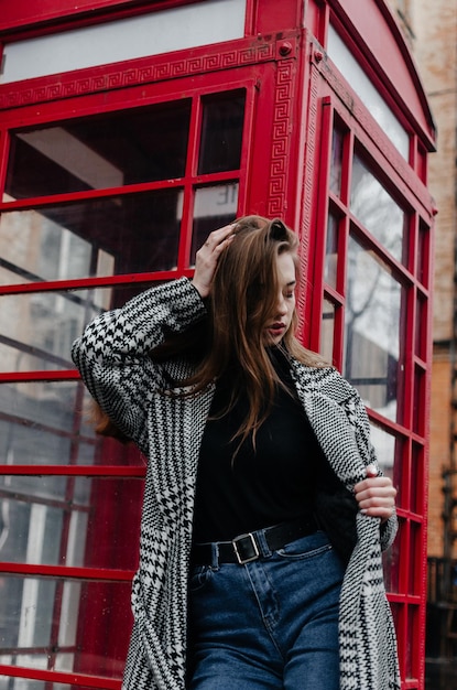 A girl in a gray coat stands by a red telephone booth