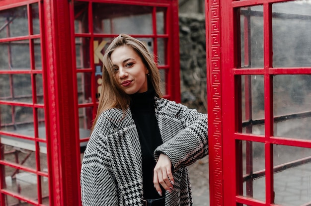 A girl in a gray coat stands by a red telephone booth