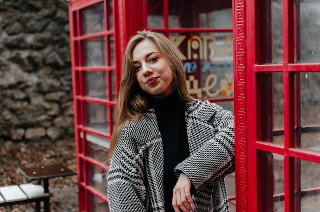 A girl in a gray coat stands by a red telephone booth