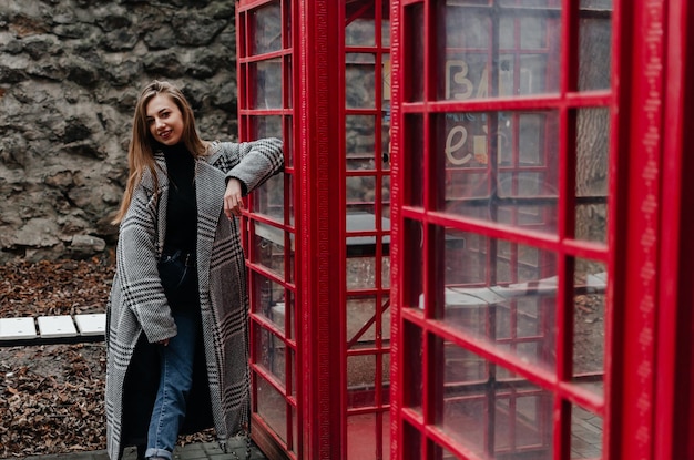 A girl in a gray coat stands by a red telephone booth