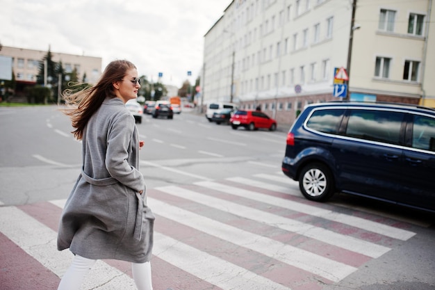 Девушка в сером пальто с солнцезащитными очками и сумочкой идет по пешеходному переходу