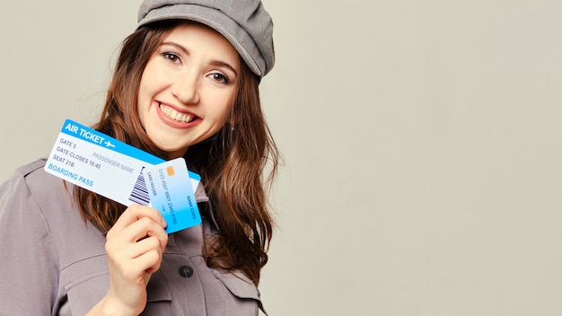 A girl in a gray dress looks at the camera and holds plane tickets with a credit card. Copy space.