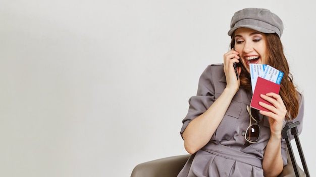 A girl in a gray dress speaks on the phone and holds plane tickets and a passport.