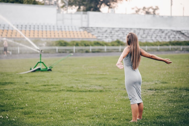 A girl in a gray dress stands with her hands apart and waits lawn sprinkler sprays water in stadium