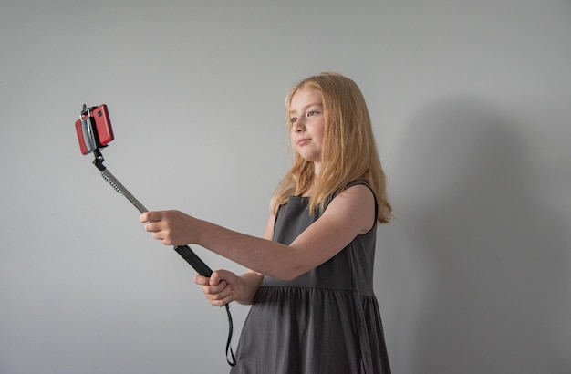 A girl in a gray dress with a selfie stick and a red phone is talking on a video call on gray background