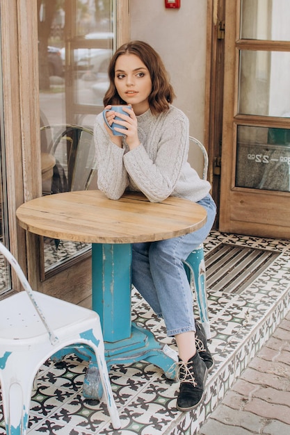 A girl in a gray sweater sits at a table in a coffee shop and drinks cocoa and enjoys the aroma