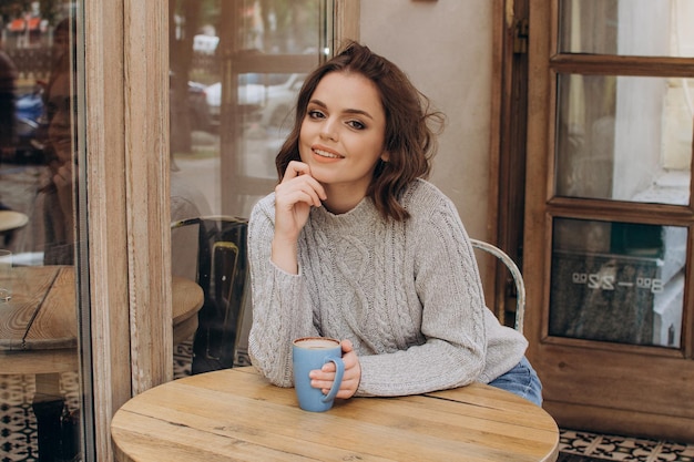 Una ragazza con un maglione grigio siede a un tavolo in una caffetteria beve cacao e sorride