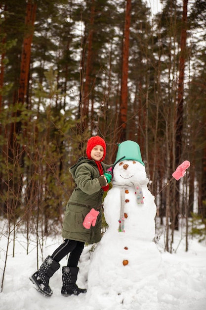 Girl in green coat in winter in forest near snowman