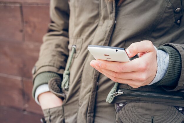 Girl in the green jacket holding a smartphone in one hand on a background of brown wall