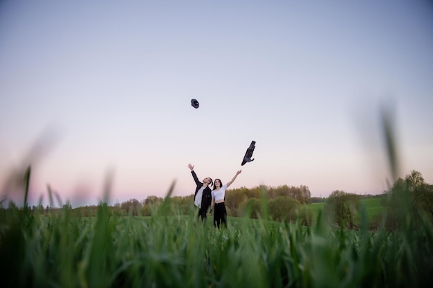 A girl and a guy are standing in a green field and tossing up a jacket and hat