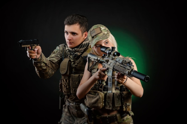 Photo a girl and a guy in military overalls pose with an airsoft gun on a dark background