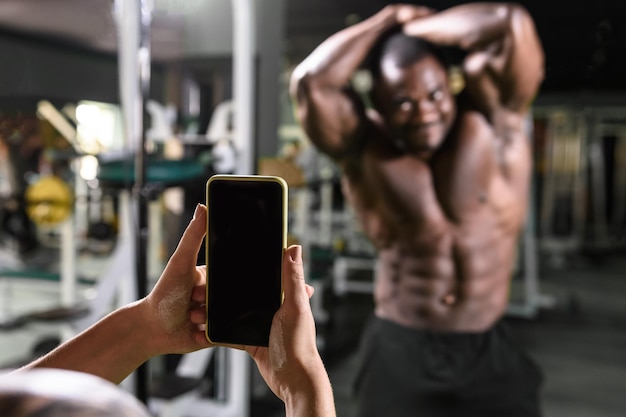 The girl in the gym takes pictures on the smartphone of an African American bodybuilder for his sports blog