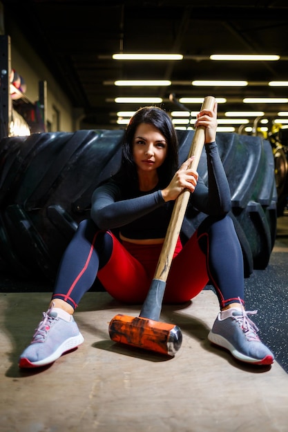 The girl in the gym with a sledgehammer in his hands sits near a big wheel