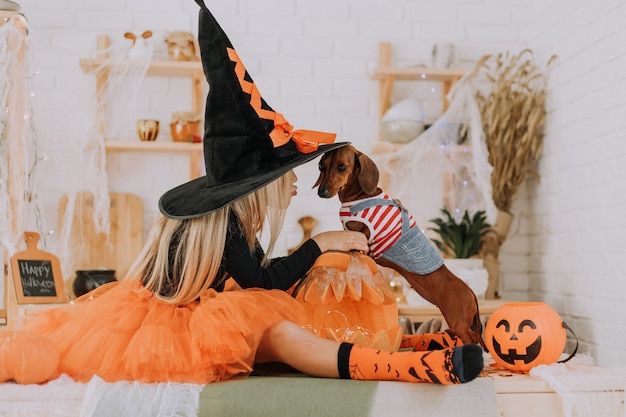 girl in Halloween witch costume and a tiny dachshund in a dog jumpsuit are sitting on floor in room