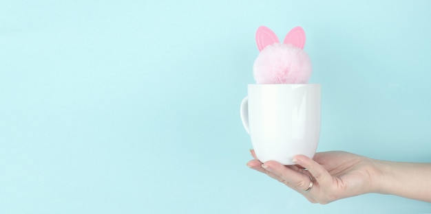 Girl hand hold morning coffee cup with pink rabbit toy inside. Copy space. Blue background.