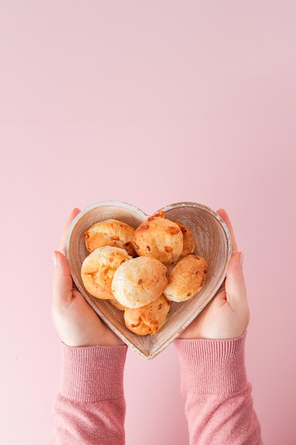 Girl hand holding Brazilian cheese bread, soft pink background, heart shaped bowl, top view vertical