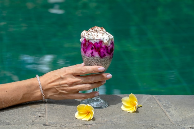 スイミングプールの水の背景にhiaシードプディングとガラスを持っている女の子の手、クローズアップ。朝食にグラスに赤いドラゴンフルーツと白いヨーグルトを添えたチアシードプリン