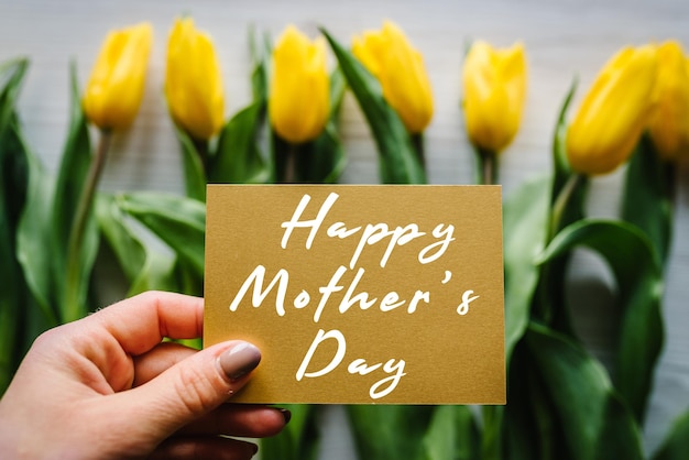 Girl hand with greeting card with text happy mother\'s day text\
sign on craft postcard on yellow tulips background greeting card\
concept sensual tender women image top view flat lay