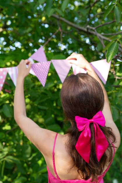 The girl hangs triangular flags of pink color against the backdrop of a green garden A girl in a pink dress and a bow on her hair Festive decor celebration concept