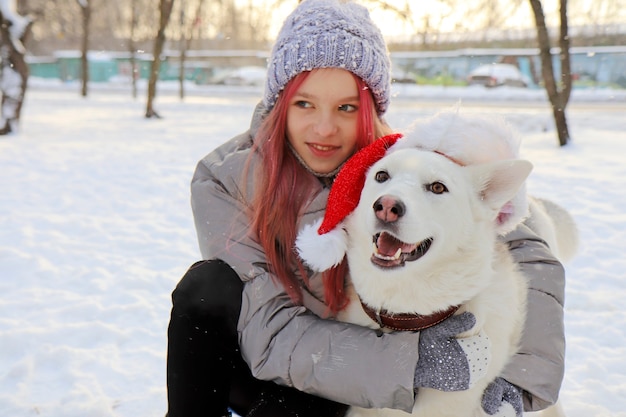 The girl happily communicates with the dog in the hat of Santa Claus in nature in winter.