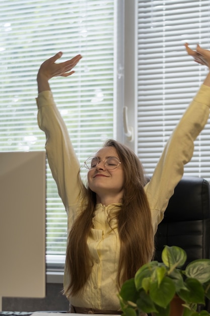 The girl happily raised her hands up and stretches while sitting at the computer. End of the working day.