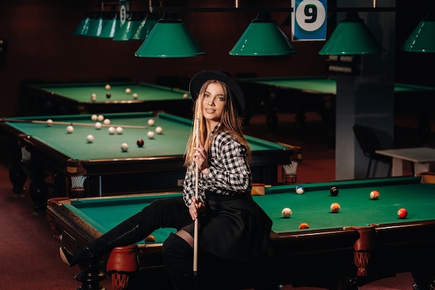 A girl in a hat in a billiard club sits on a billiard table with a cue in her hands.Playing billiards