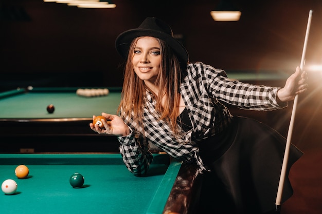 A girl in a hat in a billiard club with a cue and balls in her hands