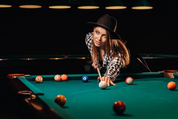 A girl in a hat in a billiard club with a cue in her hands hits a ball.Playing billiards.