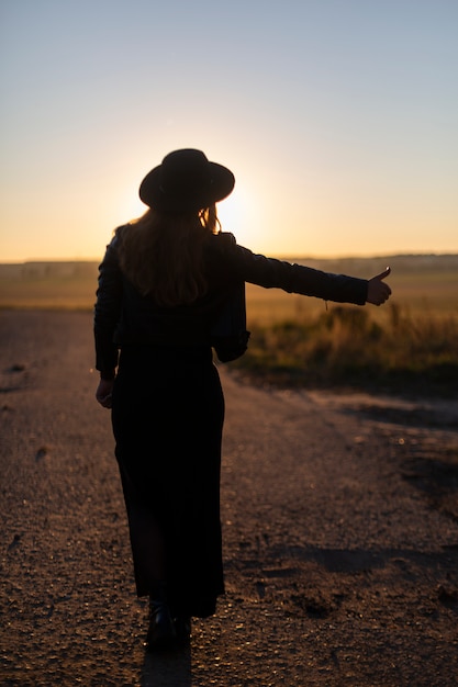 Girl in hat and dress hitchhike on desert road. Sunset. Backside. Travel concept