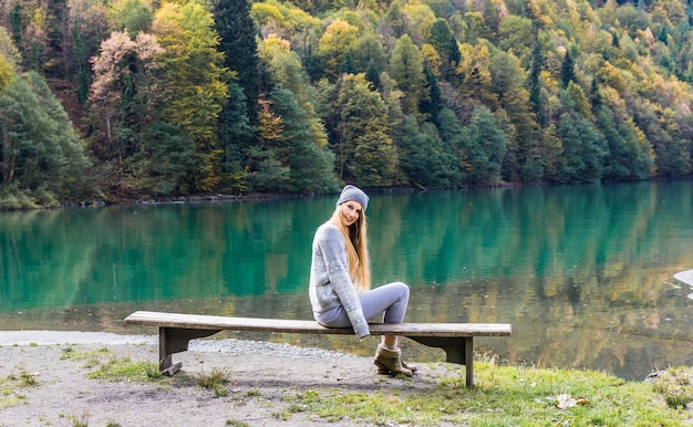 Photo a girl in a hat is sitting on a bench against the background of an autumn forest and lake