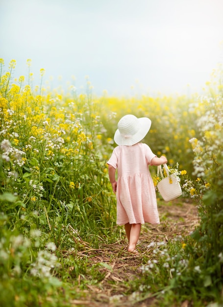 帽子をかぶった女の子が花のバスケットを持って菜種畑を歩いています
