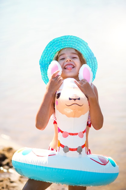 모자를 쓴 소녀는 라마 모양의 풍선 원이 있는 해안에 서 있습니다. 어린이용 풍선 알파카. 모래 바닥이 있는 바다. 해변 휴가, 수영, 선탠, 자외선 차단제.