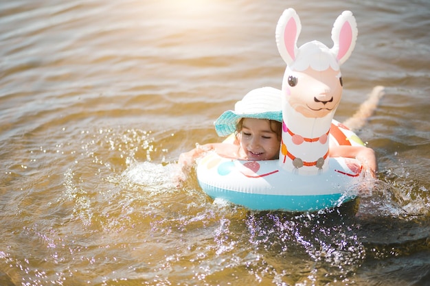 Girl in a hat it stands on the shore with inflatable circle in the shape of a lama Inflatable alpaca for a child Sea with a sandy bottom Beach holidays swimming tanning sunscreens