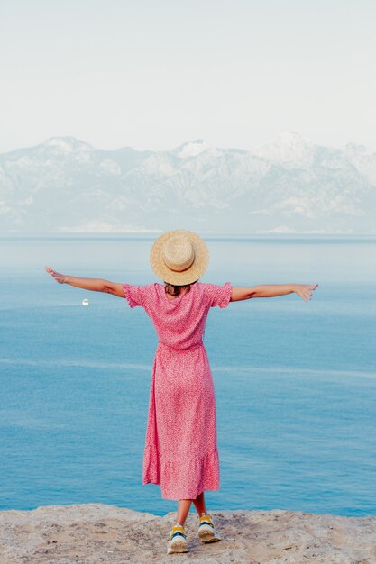 Foto ragazza con un cappello vicino al mare sul bordo di una scogliera con mare e montagne sullo sfondo