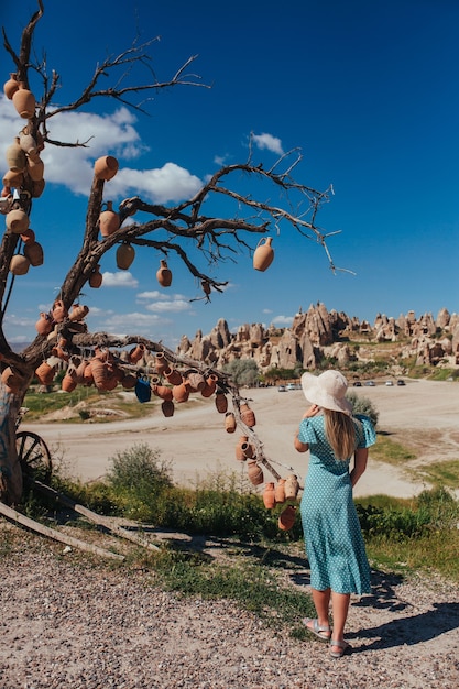 A girl in a hat and a pink dress stands near a tree hung with ceramic dishes in capadocia