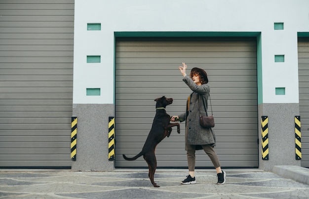 Girl in a hat plays with a dog on a background of a gray wall