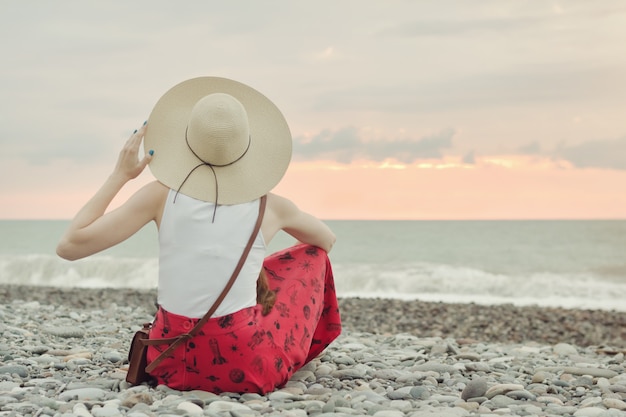 Girl in a hat sits on a pebble beach. Back view. Sunset time