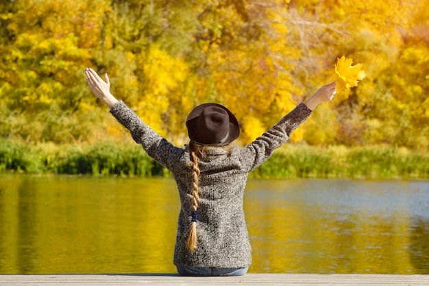 Girl in the hat sitting on the dock. Hands up. Autumn day. Back view