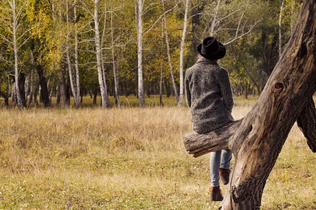 Girl in a hat sitting on a tree in the autumn forest. Back view.